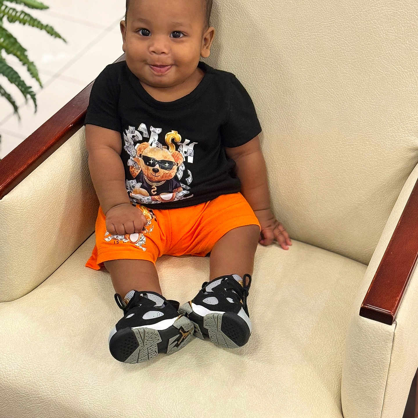 Kazi joined the competition — help win amazing prizes! armchair, baby, black_tshirt, child, cute, fashion, floor, furniture, home, indoor, leisure, orange_shorts, person, plant, portrait, sitting, smiling, sneakers, toddler, wood