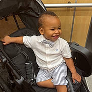 Kazi is registered to the contest to win money with this photo: toddler, child, smiling, stroller, indoor, clothing, bow_tie, shorts, shoes, happy, person, baby, seat, wheel, black, white_shirt, striped, cute, portrait, furniture