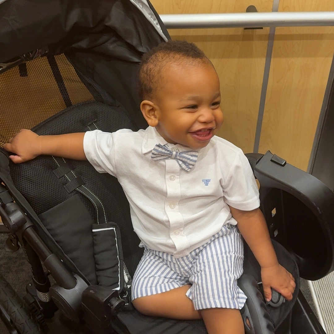 Kazi is registered to the contest to win money with this photo: toddler, child, smiling, stroller, indoor, clothing, bow_tie, shorts, shoes, happy, person, baby, seat, wheel, black, white_shirt, striped, cute, portrait, furniture