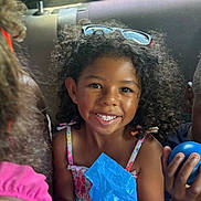 London is registered to the contest to win money with this photo: child, curly_hair, smile, sunglasses, car_seat, blue_plastic_bag, bottle_cap, clothing, backseat, happy, person, holding, toy, sunlight, portrait, casual, indoor, friends, multicolor, seat