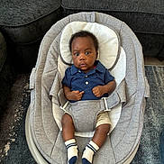 Eli is registered to the contest to win money with this photo: baby, infant, child, portrait, sitting, rocker, baby_seat, harness, blue_shirt, shorts, socks, smiling, facial_expression, indoor, living_room, sofa, carpet, home, photography, front_view