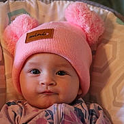 Sophia is registered to the contest to win money with this photo: baby, pink_hat, pom_poms, infant, face, child, clothing, patterned_cushion, portrait, cute, indoor, hat, knitwear, young_child, seated, expression, apparel, soft_texture, closeup, person