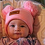 baby, pink_hat, pom_poms, infant, face, child, clothing, patterned_cushion, portrait, cute, indoor, hat, knitwear, young_child, seated, expression, apparel, soft_texture, closeup, person