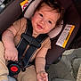 baby, child, car_seat, safety_harness, smile, face, hands, clothing, curly_hair, rosy_cheeks, infant, seatbelt, warning_label, outdoor, cute, person, closeup, toddler, seat, soft_lighting