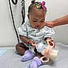 Vanessa joined the competition — help win amazing prizes! baby, infant, child, bottle, milk, hospital_gown, medical_exam_table, hand, hair_clip, socks, curly_hair, person, indoors, wall, support, cute, seated, looking_down, healthcare, caregiver