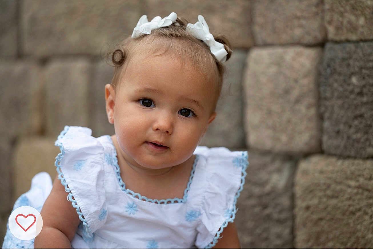 Selina joined the competition — help win amazing prizes! baby, child, girl, white_dress, bows, hair_accessories, portrait, face, cute, curious, outdoor, stone_wall, soft_lighting, closeup, infant, smiling, person, young_child, pretty, fashion