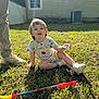 toddler, child, grass, toy, ball, golf_club, sunlight, shadow, adult_legs, shoes, outdoor, backyard, house, window, pants, shirt, footwear, play, daytime, nature
