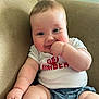 Isabella is registered to the contest to win money with this photo: baby, child, smiling, couch, seated, white_shirt, blue_shorts, hand_in_mouth, chubby, indoors, face, skin, happy, cute, person, infant, plump, short_hair, relaxed, comfortable