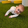 baby, child, crawling_position, curious, cute, floor, green_carpet, hands, head, indoor, infant, person, play_area, smiling, soft_lighting, tongue_out, white_clothing, wide_eyes, yellow_cushions, young_child