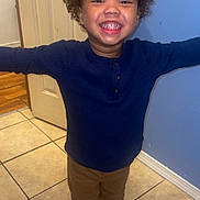 Kamari is registered to the contest to win money with this photo: child, smiling, indoor, arms_outstretched, curly_hair, navy_sweater, brown_pants, black_shoes, tile_floor, door, blue_wall, happy, person, standing, portrait, casual_clothing, young_child, full_body, home_interior, cute