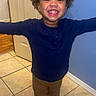 arms_outstretched, black_shoes, blue_wall, brown_pants, casual_clothing, child, curly_hair, cute, door, full_body, happy, home_interior, indoor, navy_sweater, person, portrait, smiling, standing, tile_floor, young_child