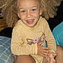 Denali is registered to the contest to win money with this photo: bed, bedding, blue_pillow, casual, child, cozy, curly_hair, cute, face, hand, happy, indoor, patterned_blanket, person, portrait, sitting, skin, smiling, yellow_sweater, young