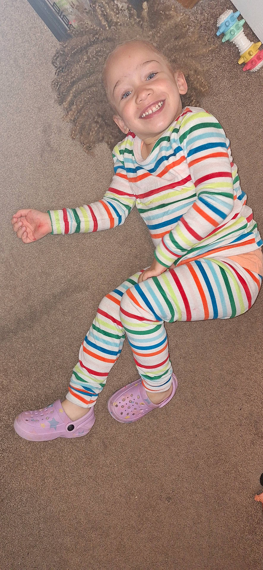 bright, carpet, casual, cheerful, child, curly_hair, cute, footwear, happy, indoor, joyful, lying_down, pajamas, person, playful, purple_crocs, relaxed, smiling, striped_clothing, young