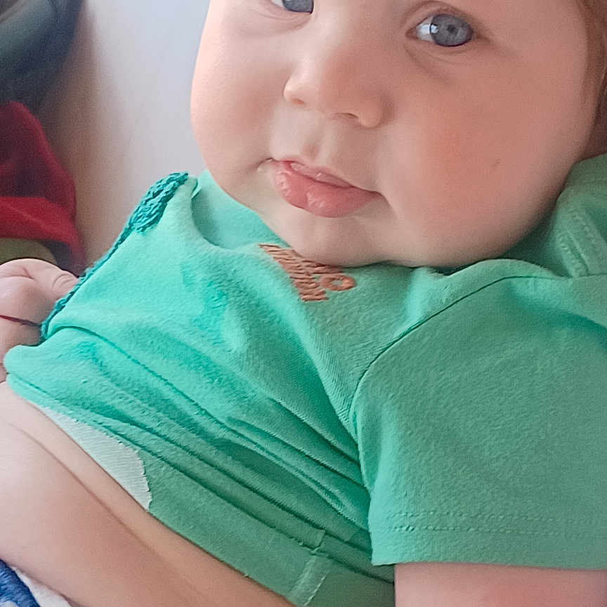 Roan is registered to the contest to win money with this photo: baby, child, thumbs_up, green_shirt, blue_eyes, cute, infant, portrait, hand_gesture, curly_hair, indoor, skin, face, chubby, expression, person, sitting, casual_clothing, diaper, light