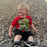 Ace is registered to the contest to win money with this photo: toddler, child, smiling, red_shirt, dinosaur_graphic, rocks, pile_of_rocks, outdoor, blonde_hair, black_shorts, black_sneakers, casual_clothing, happy, sitting, playful, daylight, nature, ground, person, portrait