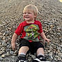toddler, child, smiling, red_shirt, dinosaur_graphic, rocks, pile_of_rocks, outdoor, blonde_hair, black_shorts, black_sneakers, casual_clothing, happy, sitting, playful, daylight, nature, ground, person, portrait