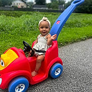 Kaylani is registered to the contest to win money with this photo: toddler, child, toy_car, red, blue, headband, outdoor, grass, road, smiling, happy, summer, play, vehicle, person, nature, greenery, daytime, park, casual