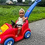 toddler, child, toy_car, red, blue, headband, outdoor, grass, road, smiling, happy, summer, play, vehicle, person, nature, greenery, daytime, park, casual