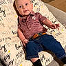 baby, infant, child, polo_shirt, jeans, socks, cushion, couch, indoor, smile, curious, face, person, clothing, text, home, comfortable, seated, reaching, casual