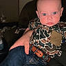 Cooper is registered to the contest to win money with this photo: baby, child, person, infant, camo_shirt, blue_jeans, belt, hand, indoor, portrait, facial_expression, seated, casual_clothing, adult_hand, furniture, dim_lighting, focused, closeup, human, cute