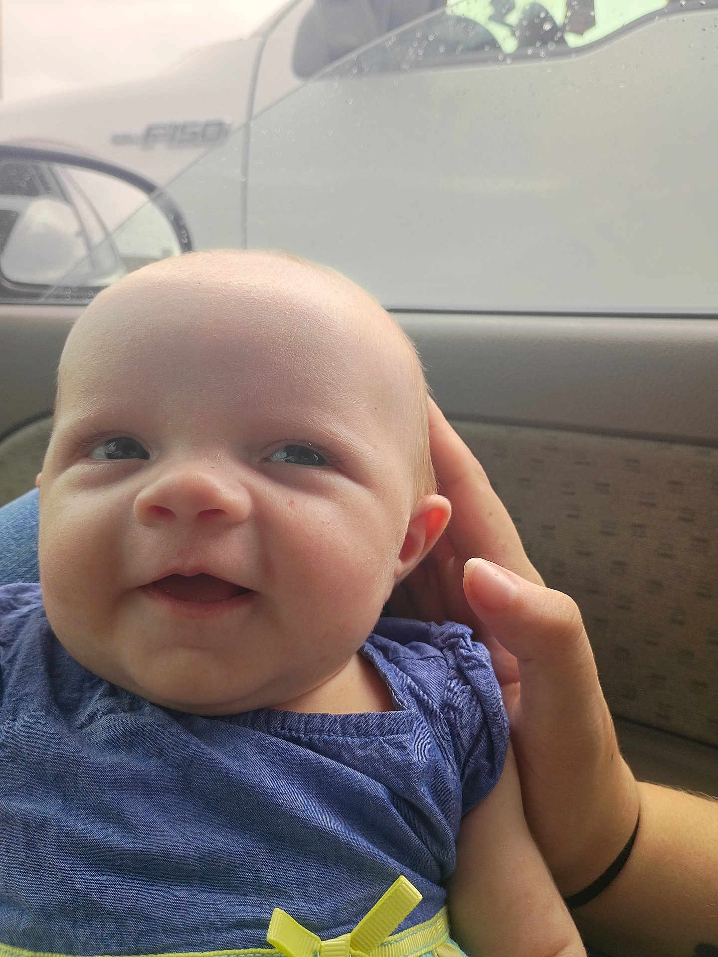 Harmony is registered to the contest to win money with this photo: baby, child, smile, face, hand, blue_dress, yellow_bow, car_interior, window, rain_drops, person, skin, cute, infant, portrait, closeup, seat, clothing, human, happy