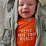 baby, smiling, hoodie, orange_shirt, text, blanket, infant, cute, child, happy, face, apparel, clothing, portrait, indoor, baby_garment, soft_texture, young_child, casual_wear, relaxed