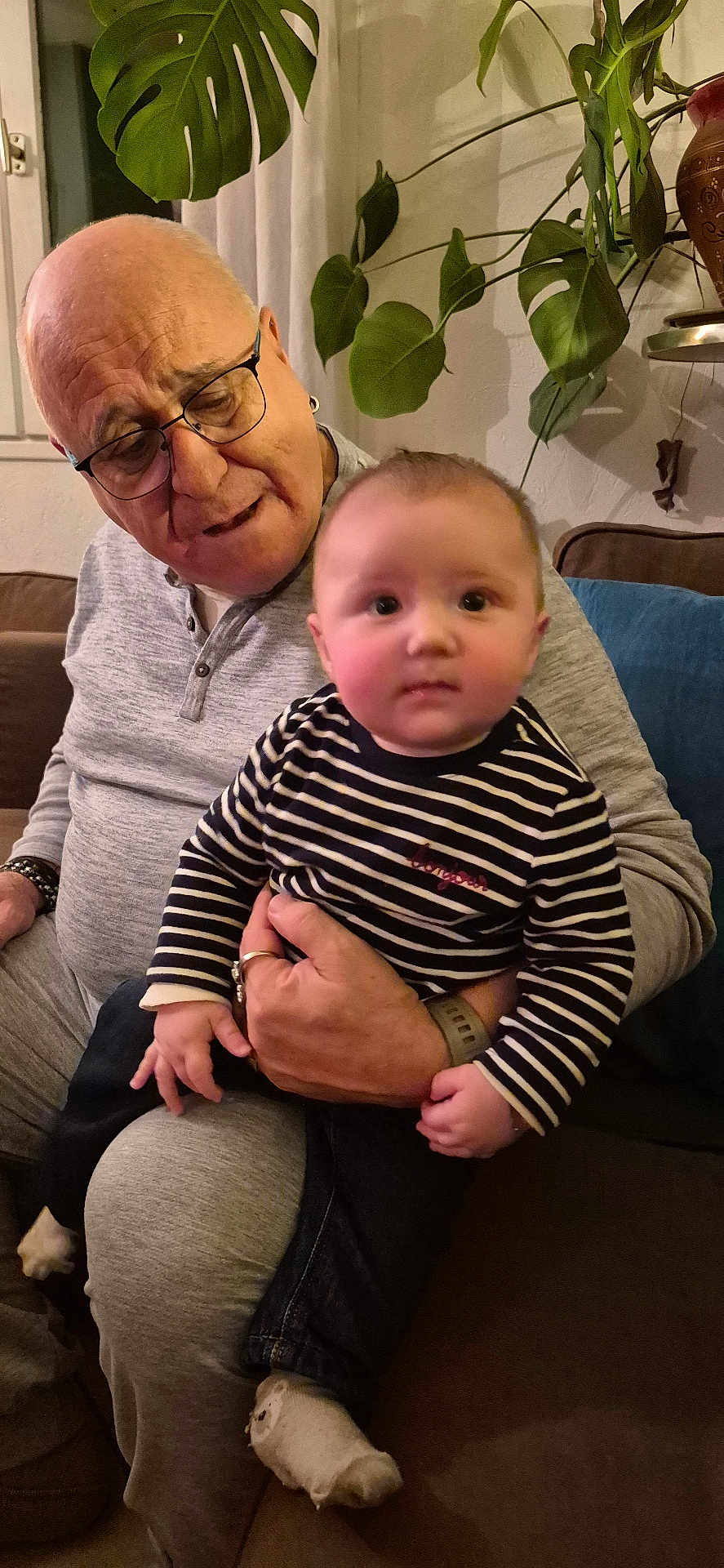 Temae a rejoint le concours — aidez-le/la à gagner de superbes lots ! elderly_man, baby, child, striped_shirt, glasses, couch, indoor, plants, home_decor, cozy, family, holding, socks, portrait, seated, casual_clothing, living_room, affection, tender_moment, people
