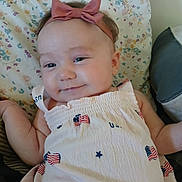 Belladonna is registered to the contest to win money with this photo: baby, infant, headband, bow, dress, usa_flag, pattern, floral_blanket, smile, face, skin, arm, lying_down, cute, child, indoors, fabric, person, apparel, textile