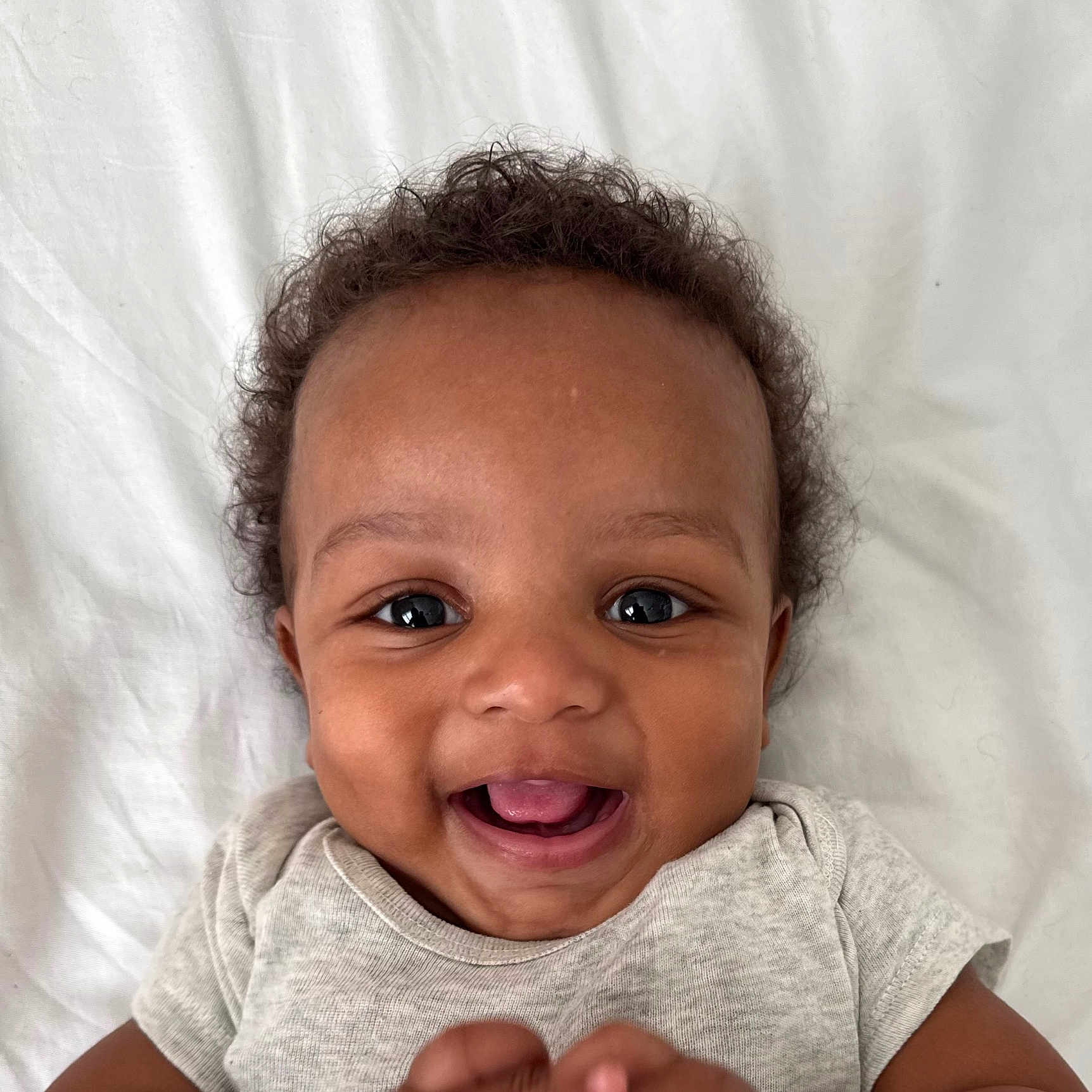 Eziah is registered to the contest to win money with this photo: baby, smiling, happy, infant, face, cute, child, closeup, portrait, hands, clothing, curly_hair, lying_down, light_grey_shirt, white_background, skin, young_child, joyful, playful, expressive