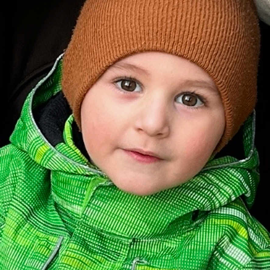 Nicolas is registered to the contest to win money with this photo: beanie, brown_hat, cheeks, child, closeup, cute, eyebrows, eyes, face, green_jacket, hood, outfit, portrait, seat, smile, stroller, toddler, warm_clothing, winter, winter_jacket