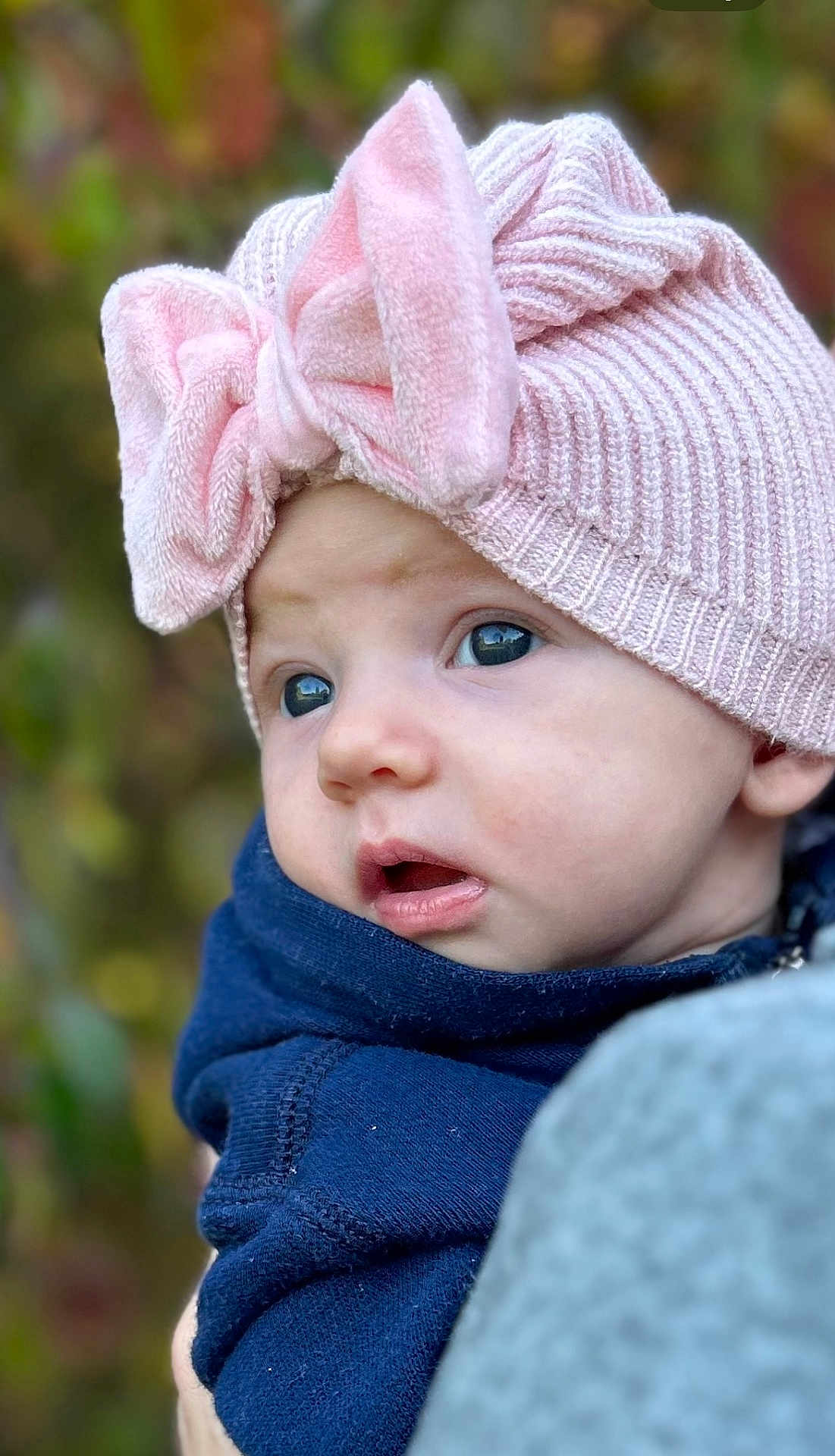 Amelia is registered to the contest to win money with this photo: baby, child, face, hat, pink, bow, knit, blue_clothing, portrait, outdoor, curious, closeup, infant, soft_focus, cute, warm_clothing, person, headwear, expression, fall_background