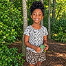Ava is registered to the contest to win money with this photo: child, curly_hair, daylight, floral_shirt, forest, full_body, girl, greenery, happy, leaf, leopard_print, nature, outdoors, portrait, shorts, smiling, sneakers, standing, trees, woods