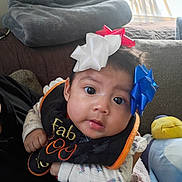 Camila is registered to the contest to win money with this photo: baby, infant, face, eyes, bow, headwear, bib, clothing, hand, blanket, couch, indoor, soft_toys, plush, person, cute, expression, portrait, child, cozy