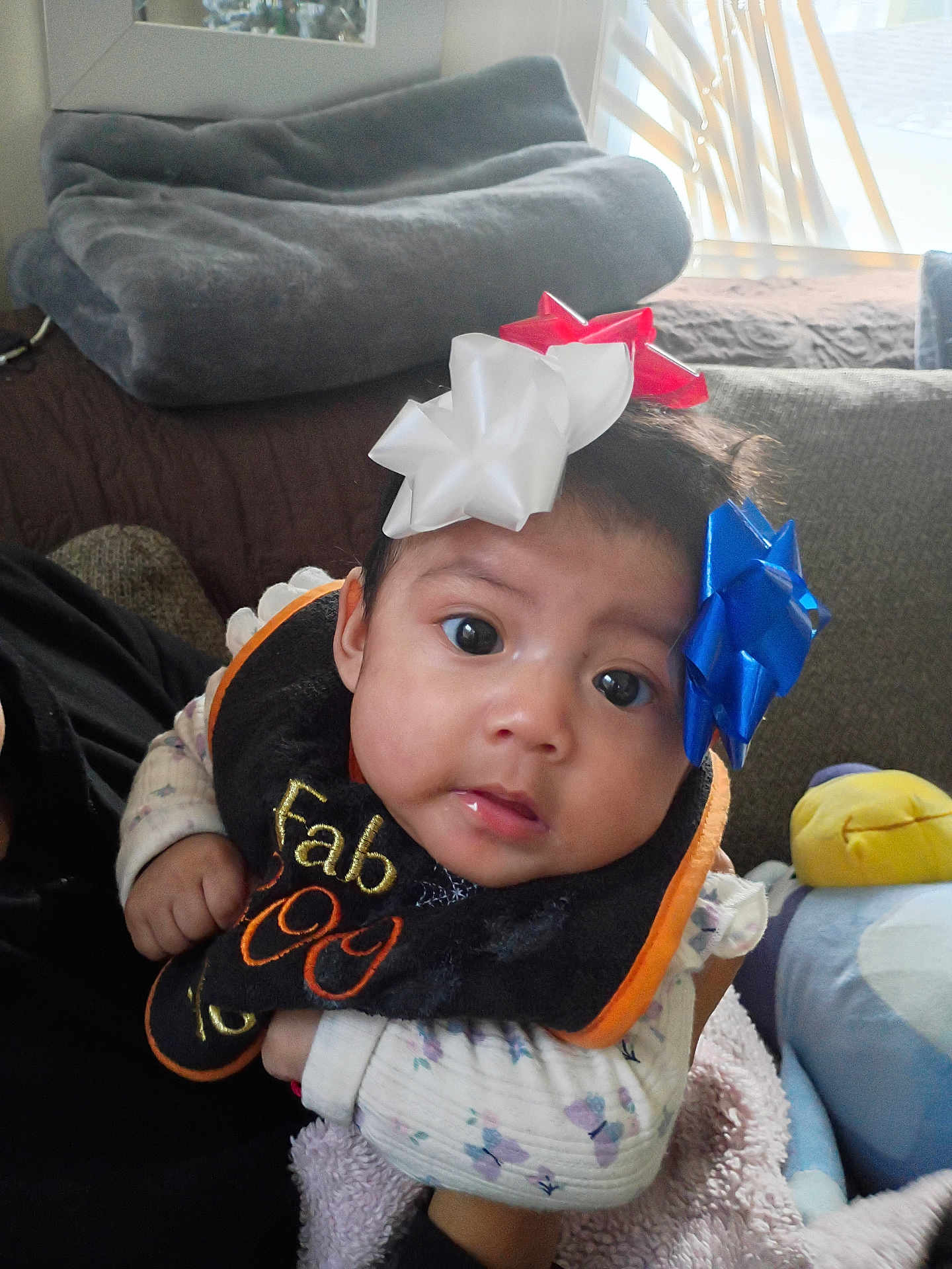 Camila is registered to the contest to win money with this photo: baby, infant, face, eyes, bow, headwear, bib, clothing, hand, blanket, couch, indoor, soft_toys, plush, person, cute, expression, portrait, child, cozy