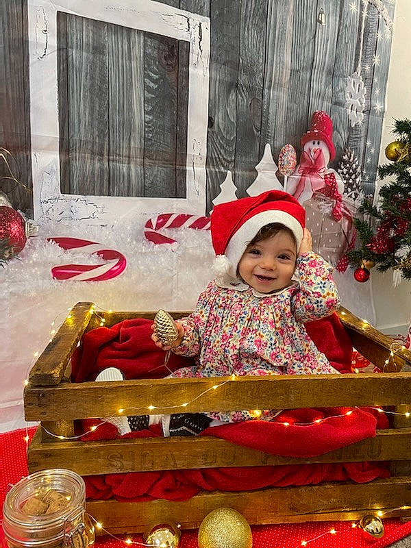 Lina a rejoint le concours — aidez-le/la à gagner de superbes lots ! baby, baby_toddler_clothing, child, christmas, christmas_decoration, christmas_eve, christmas_ornament, decoration, event, happy, hat, headwear, holiday, joy, ornament, person, red, room, sitting, smile