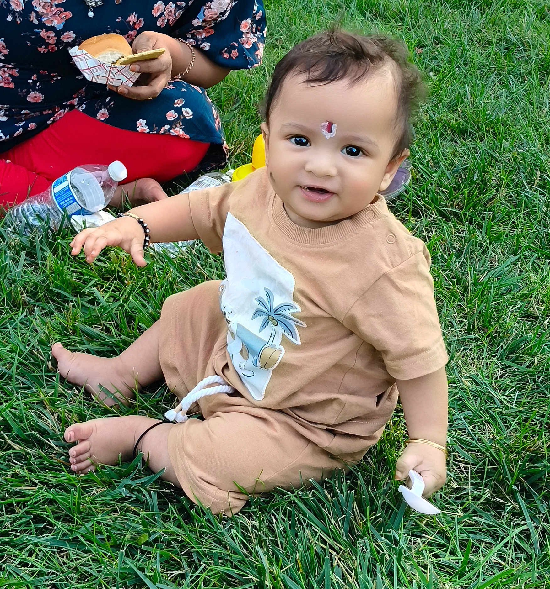 Praneeth Thunugunta is registered to the contest to win money with this photo: baby, child, grass, outdoor, pacifier, beige_outfit, sitting, smiling, food, sandwich, adult, floral_dress, red_leggings, picnic, bracelet, bracelet_black, barefoot, happy, nature, casual