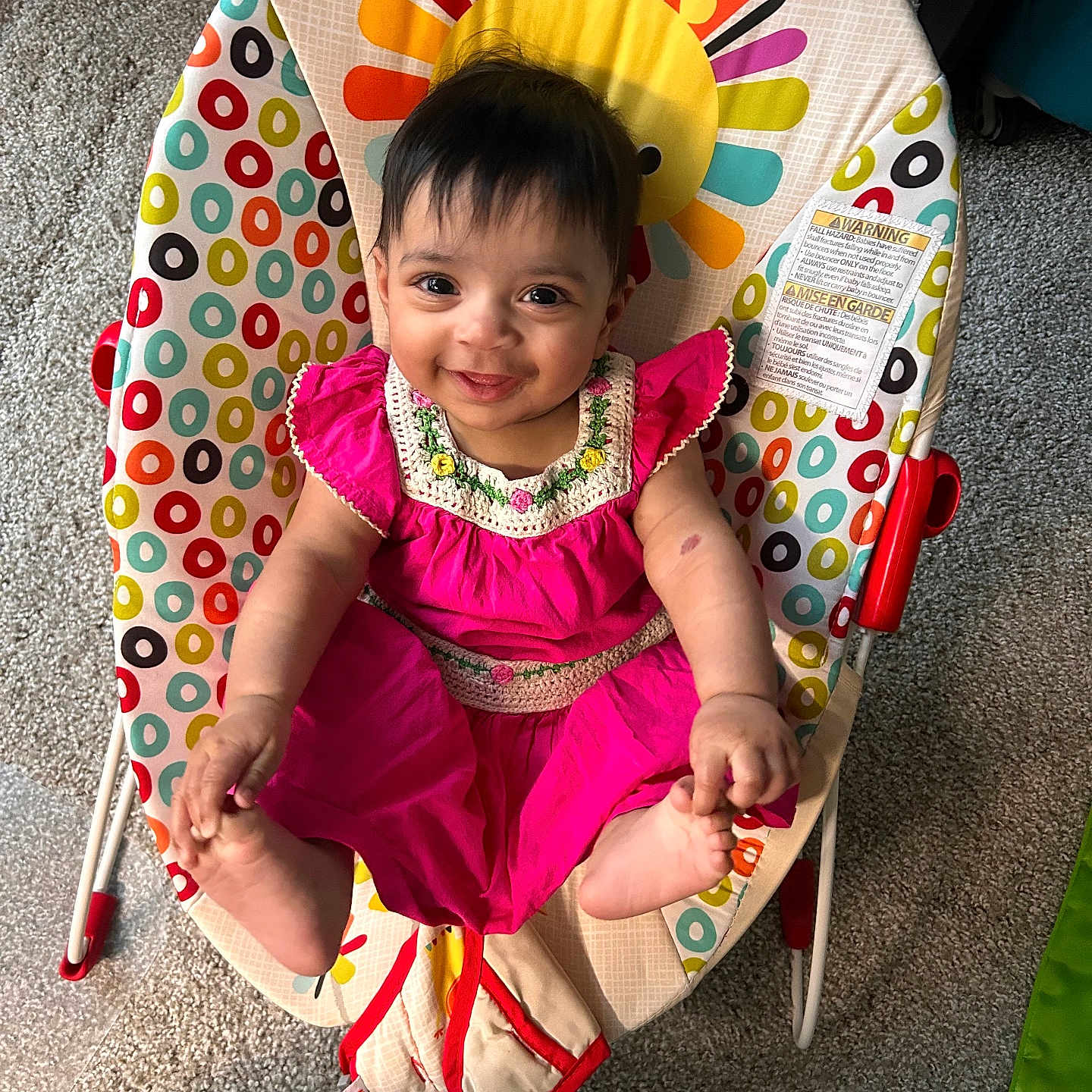 Eliza is registered to the contest to win money with this photo: baby, baby_chair, carpet, child, colorful, cute, face, feet, hair, hand, happy, indoor, infant, pattern, person, pink_dress, seat, smiling, sun_design, toy