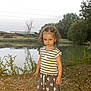 Mya a rejoint le concours — aidez-le/la à gagner de superbes lots ! child, girl, curly_hair, striped_shirt, skirt, pond, water, trees, grass, leaves, outdoor, nature, daylight, shoes, person, young_child, summer, casual_clothing, reflection, bushes