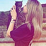 woman, cat, outdoor, sky, nature, blonde_hair, holding, affection, tabby_cat, casual_clothing, watch, bracelet, wall, greenery, summer, daylight, portrait, side_view, smile, human_pet_interaction