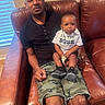 man, baby, child, couch, leather, tshirt, pacifier, window, blinds, camouflage_shorts, indoor, person, seated, living_room, flooring, expression, holding, family, casual, portrait