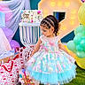 child, toddler, birthday, birthday_cake, party, dress, pastel, balloons, presents, gift_bags, smiling, portrait, standing, girl, curly_hair, braids, outdoor, colorful, decorations, number_backdrop