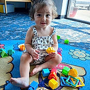 Sophia is registered to the contest to win money with this photo: toddler, child, curly_hair, play_mat, toys, plastic_toys, plush_giraffe, indoor, natural_light, window, crib, colorful, smiling, sitting, cross_legged, casual_clothing, playtime, happy, young_child, floor