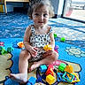 toddler, child, curly_hair, play_mat, toys, plastic_toys, plush_giraffe, indoor, natural_light, window, crib, colorful, smiling, sitting, cross_legged, casual_clothing, playtime, happy, young_child, floor