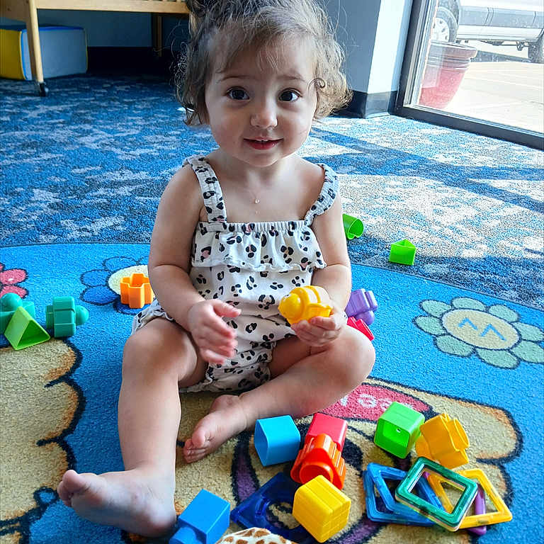 Sophia is registered to the contest to win money with this photo: toddler, child, curly_hair, play_mat, toys, plastic_toys, plush_giraffe, indoor, natural_light, window, crib, colorful, smiling, sitting, cross_legged, casual_clothing, playtime, happy, young_child, floor