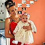 Abby a rejoint le concours — aidez-le/la à gagner de superbes lots ! woman, baby, smile, red_dress, white_dress, headband, tattoo, photographs, orange_wall, indoor, holding, happy, portrait, person, home, decor, hand, face, clothing, room