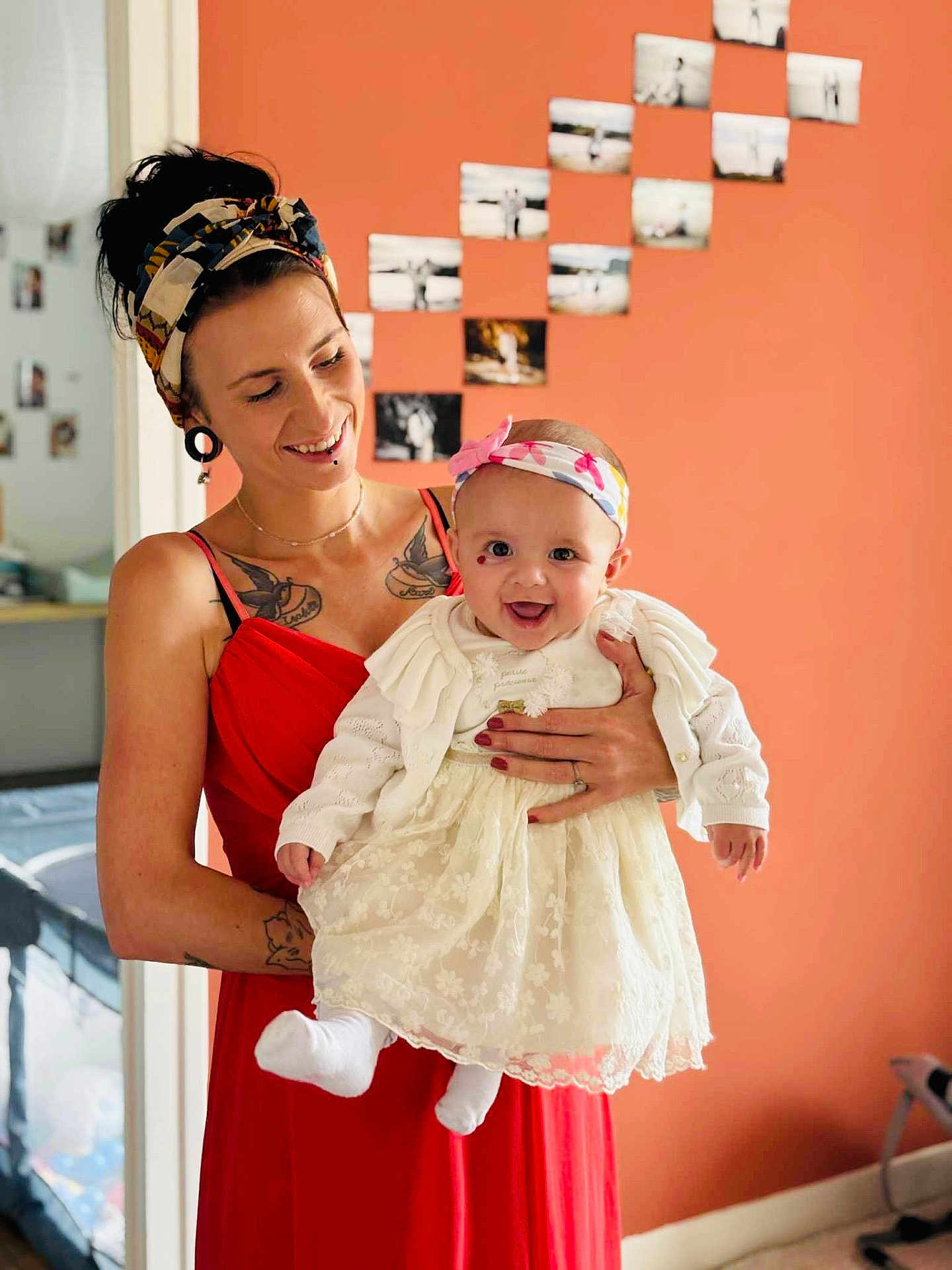 Abby a rejoint le concours — aidez-le/la à gagner de superbes lots ! woman, baby, smile, red_dress, white_dress, headband, tattoo, photographs, orange_wall, indoor, holding, happy, portrait, person, home, decor, hand, face, clothing, room