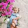 Victoire participe au concours pour gagner de l'argent avec cette photo : toddler, child, blue_romper, flowering_bush, pink_flowers, adult_hands, stone_wall, window, outdoor, daylight, curious_expression, playful, holding, skin, short_hair, footwear, nature, person, portrait, green_leaves