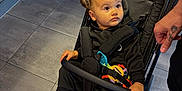 Skylar joined the competition — help win amazing prizes! toddler, child, stroller, indoor, flooring, tile, black_clothing, pink_shoes, hand, tattoo, safety_bar, curly_hair, baby_toy, straps, seated, looking_away, cabinet, adult, footwear, expression