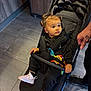 toddler, child, stroller, indoor, flooring, tile, black_clothing, pink_shoes, hand, tattoo, safety_bar, curly_hair, baby_toy, straps, seated, looking_away, cabinet, adult, footwear, expression