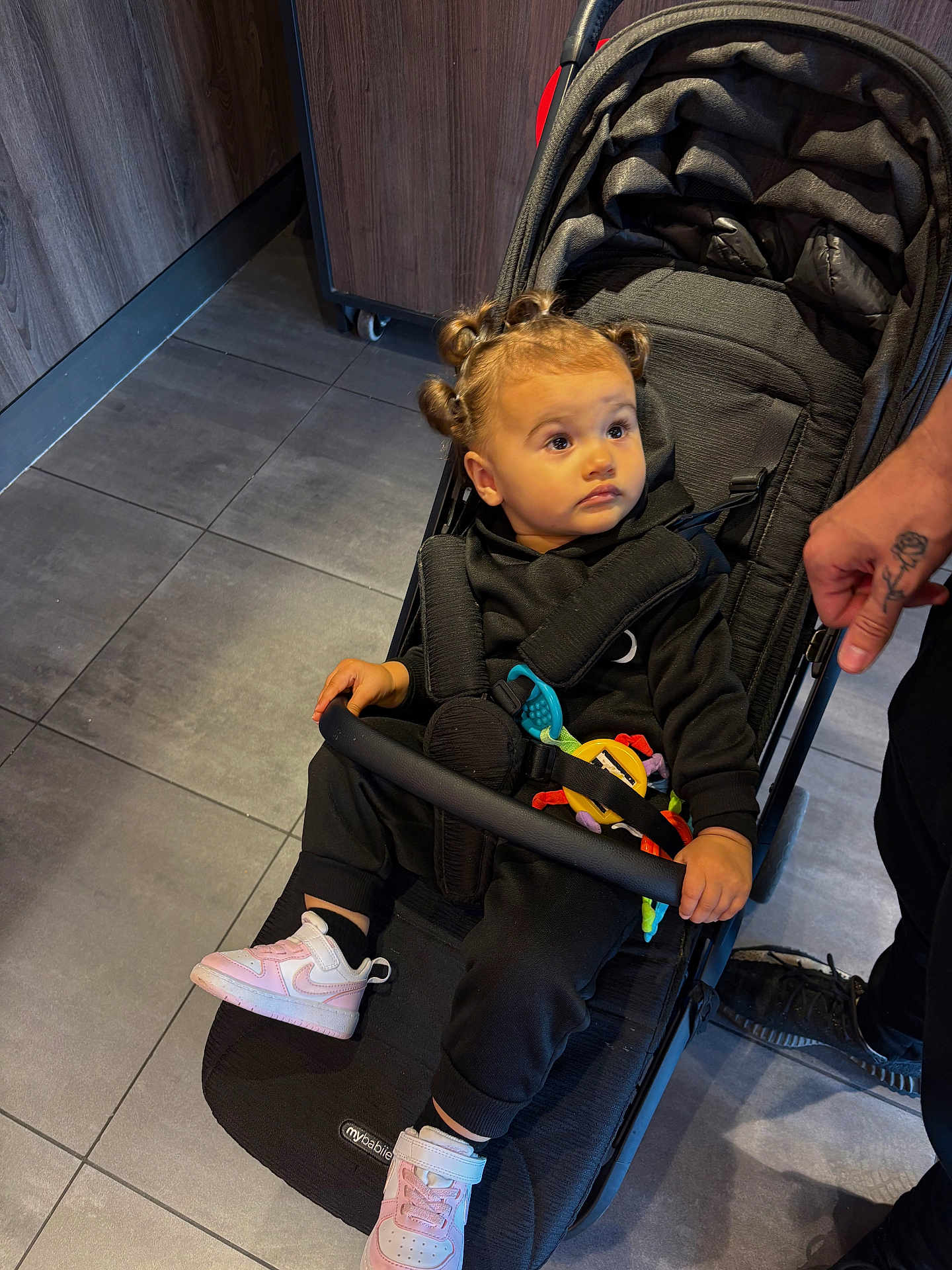 Skylar joined the competition — help win amazing prizes! toddler, child, stroller, indoor, flooring, tile, black_clothing, pink_shoes, hand, tattoo, safety_bar, curly_hair, baby_toy, straps, seated, looking_away, cabinet, adult, footwear, expression